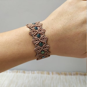May include: A macrame bracelet featuring a floral design with brown cord and teal beads. The bracelet is worn on a wrist and has a woven pattern. The bracelet is approximately 1 inch wide.