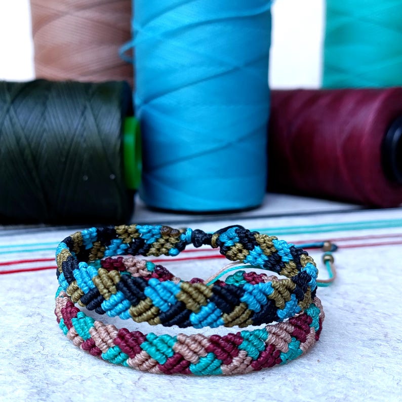 K&ouml;nnte beinhalten: Zwei Freundschaftsarmb&auml;nder aus buntem Stickgarn. Die Armb&auml;nder sind im Chevron-Muster mit Blaut&ouml;nen, Gr&uuml;n, Schwarz, Braun und Rot gewebt.