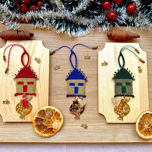 Pu&ograve; includere: Tre addobbi natalizi fatti a mano a forma di case con sfondo dorato e una croce rossa, blu e verde. Ogni addobbo ha un ciondolo a forma di melograno dorato.