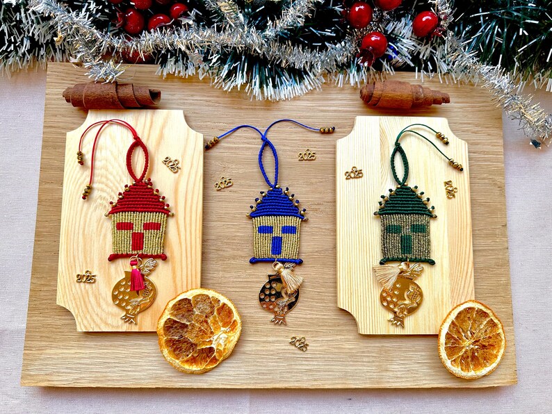 Pu&ograve; includere: Tre addobbi natalizi fatti a mano a forma di case con sfondo dorato e una croce rossa, blu e verde. Ogni addobbo ha un ciondolo a forma di melograno dorato.