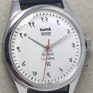 May include: Silver-toned wristwatch with a white face and black numerals. The watch features a black leather band and a red second hand. The watch face includes text in a foreign language.