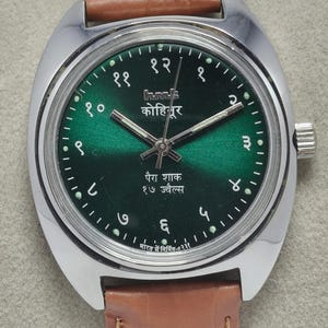 May include: A vintage wristwatch with a green dial and silver-tone case. The watch features Arabic numerals and hands, with text in an Indian language. It has a brown leather strap.