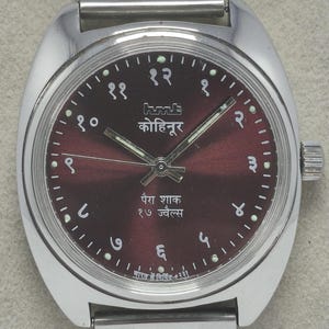 May include: A silver-toned wristwatch with a burgundy face. The watch features Arabic numerals and text in a foreign language. The watch band is also silver-toned and made of linked metal. The watch has a classic, vintage design.