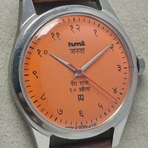 May include: Close-up of a vintage wristwatch with an orange face and a brown leather band. The watch has silver casing and features Hindi numerals and text. The watch hands are silver and red.
