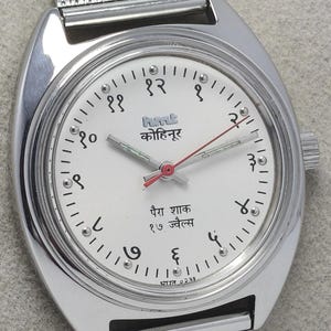 May include: Silver-toned wristwatch with a white face featuring black numerals and Hindi script. The watch has a mesh band and a red second hand. The watch face includes the text "hmk कोहिनूर पैरा शाक १७ ज्वैल्स भारत ०२३१".