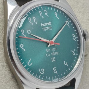 May include: Close-up of a silver wristwatch with a teal face and a green leather strap. The watch face features Arabic numerals and text in a foreign language. The watch hands are silver and red.
