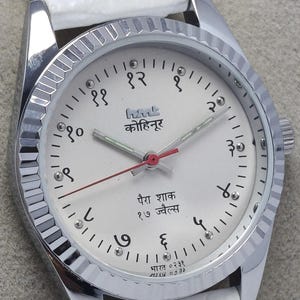 May include: A silver-toned wristwatch with a white face and a white leather strap. The watch face features black numerals, Hindi script, and red and silver hands. The watch has a fluted bezel.