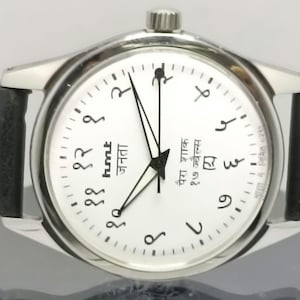 May include: A silver wristwatch with a white face and black hands. The watch has a black leather band and features Arabic numerals and the text "hmt जनता पैरा शाक १७ ज्वेल्स".