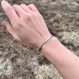 May include: A silver bangle bracelet on a person's wrist. The bracelet is simple and has a smooth, polished finish.