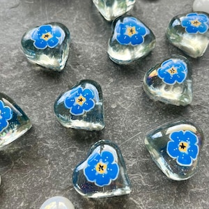 May include: A collection of clear glass heart-shaped cabochons, each featuring a painted blue five-petalled flower with a yellow centre. The hearts are interspersed with small, smooth, opaque white glass spheres on a dark grey textured surface.