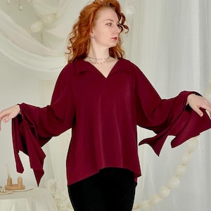 May include: A burgundy blouse with wide, flowing sleeves and a V-neck. The blouse is paired with black pants. The background features white curtains and a decorative chandelier, creating a soft, elegant aesthetic.