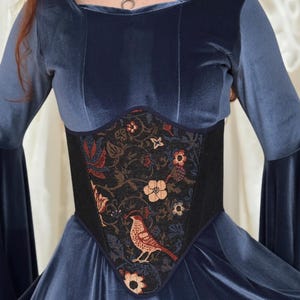 May include: A blue velvet long-sleeved dress featuring a central corset-style panel. The panel is black with a woven pattern of colorful flowers and a bird. A silver-toned chainmail-style necklace with a crescent moon pendant is worn at the neck.