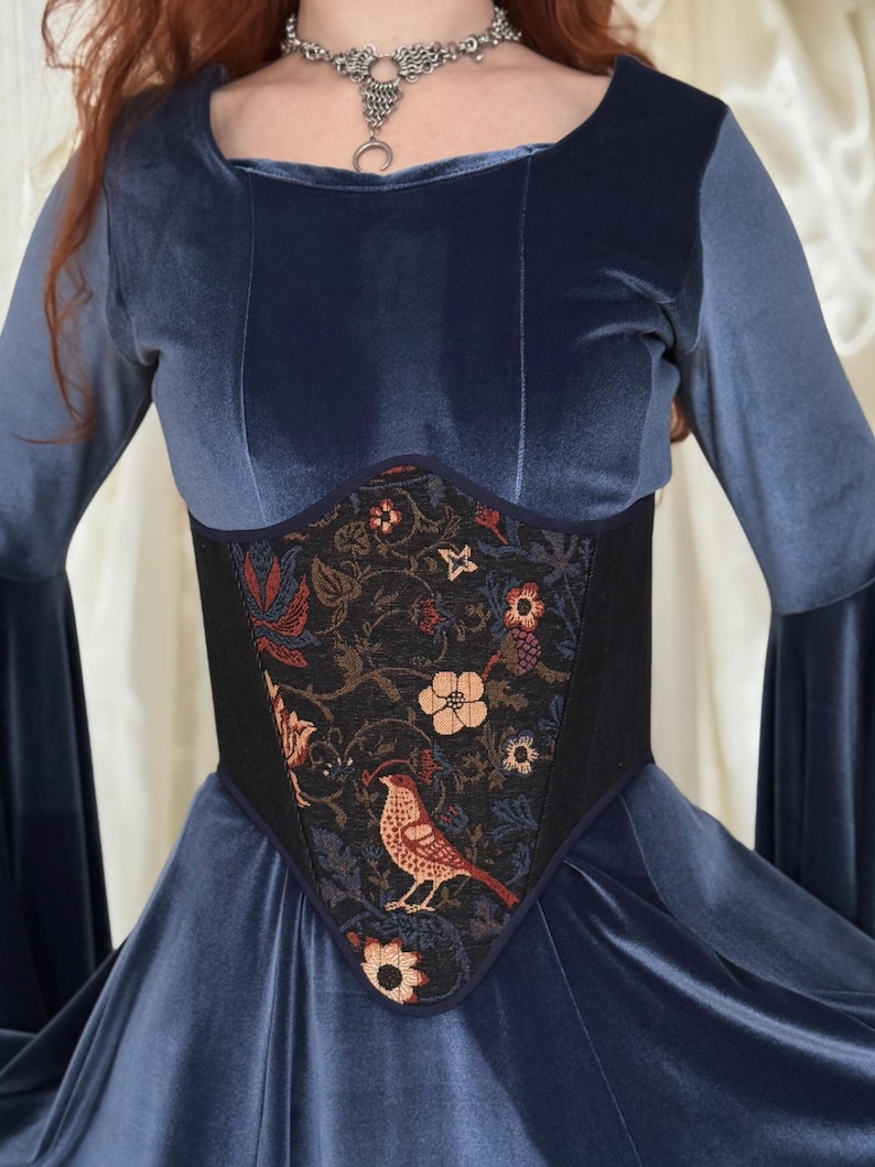 May include: A blue velvet long-sleeved dress featuring a central corset-style panel. The panel is black with a woven pattern of colorful flowers and a bird. A silver-toned chainmail-style necklace with a crescent moon pendant is worn at the neck.