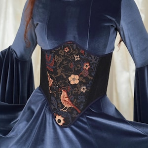 May include: A long-sleeved blue velvet dress featuring a fitted bodice with a central black corset-style panel. The panel is decorated with a detailed woven pattern of colorful flowers, vines, and a bird. A silver chain necklace is worn at the neck.