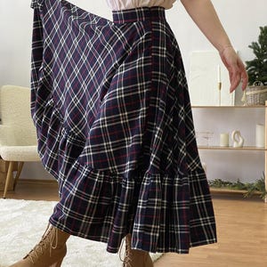 Puede incluir: Falda larga y fluida con un estampado de cuadros azul marino, rojo y blanco. La falda tiene un dobladillo con volantes y se combina con botines color canela. La falda se levanta para mostrar su forma completa.