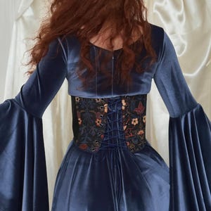 May include: A dark blue velvet dress with long flared sleeves and a fitted bodice. A black corset-style waist cincher with a multi-colored floral and botanical print is worn over the dress, featuring a dark blue lace-up closure at the back.