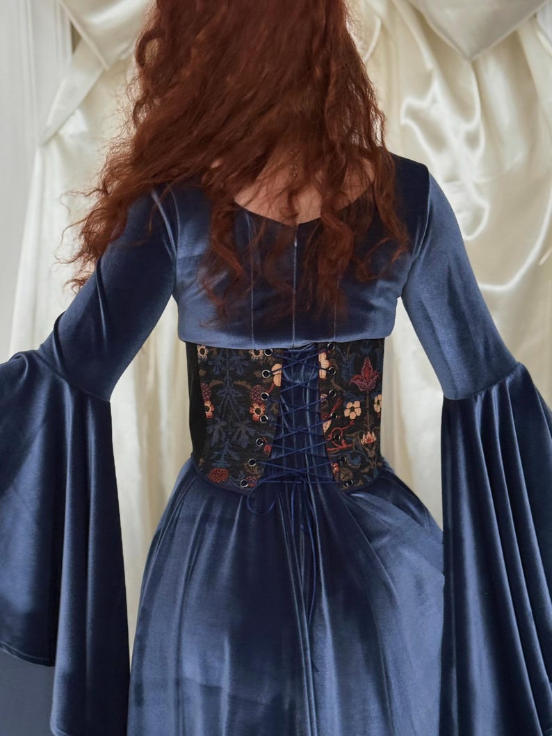May include: A dark blue velvet dress with long flared sleeves and a fitted bodice. A black corset-style waist cincher with a multi-colored floral and botanical print is worn over the dress, featuring a dark blue lace-up closure at the back.
