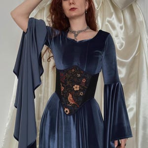 May include: A long-sleeved blue velvet dress with wide flared sleeves and a black corset-style waist panel featuring an embroidered pattern of flowers and a bird. The dress has a scoop neckline and a floor-length skirt with a soft, reflective finish.