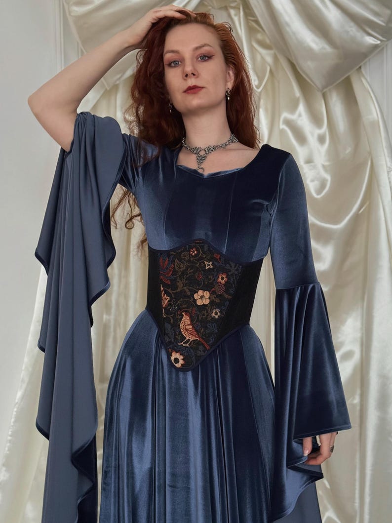 May include: A long-sleeved blue velvet dress with wide flared sleeves and a black corset-style waist panel featuring an embroidered pattern of flowers and a bird. The dress has a scoop neckline and a floor-length skirt with a soft, reflective finish.