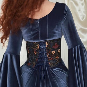 May include: A navy blue velvet dress featuring long bell sleeves and a scoop neckline. The waist is cinched with a black corset-style belt adorned with a colorful floral pattern and a central lace-up closure with matching navy blue cord lacing.