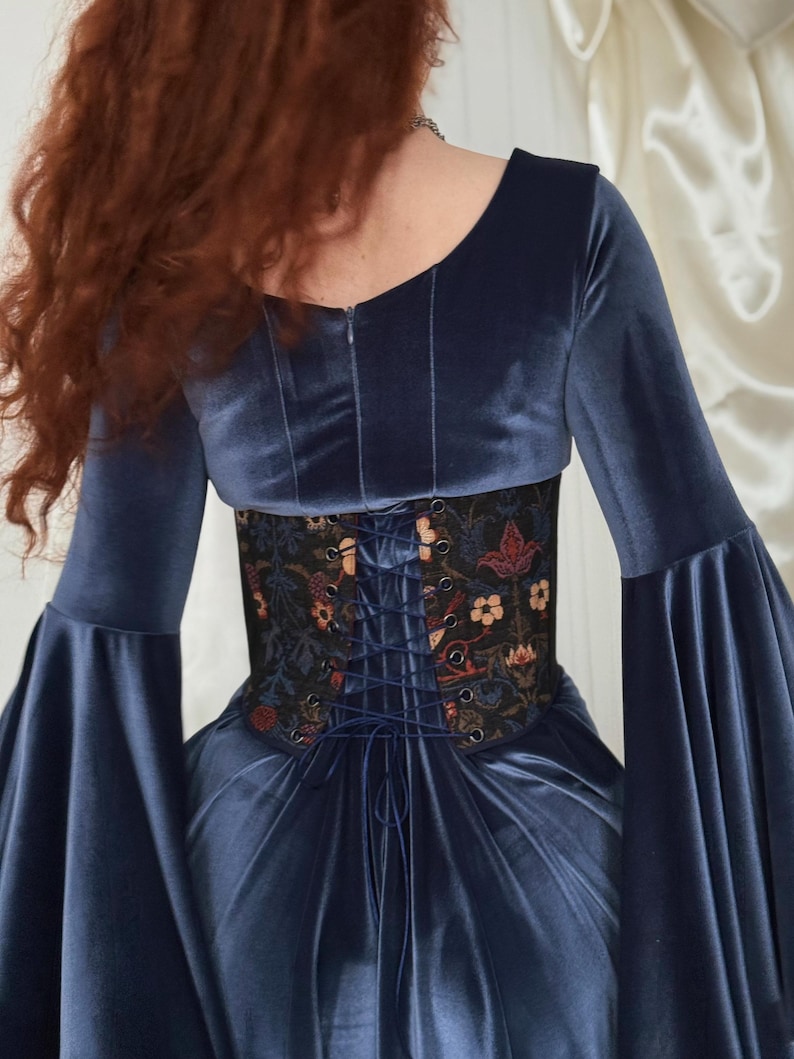 May include: A navy blue velvet dress featuring long bell sleeves and a scoop neckline. The waist is cinched with a black corset-style belt adorned with a colorful floral pattern and a central lace-up closure with matching navy blue cord lacing.