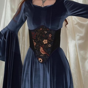 May include: A long-sleeved navy blue velvet dress with wide flared sleeves and a square neckline. The dress features a black corset-style waist panel adorned with a multi-colored floral and bird embroidery pattern. A silver chain necklace is worn.