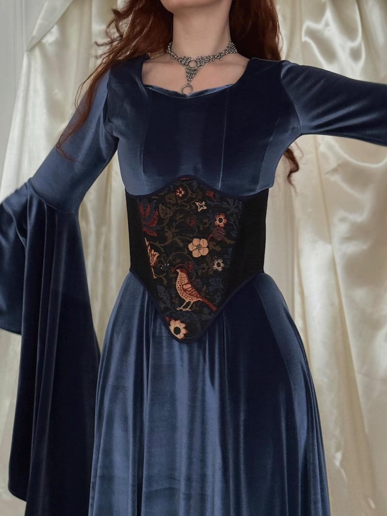 May include: A long-sleeved navy blue velvet dress with wide flared sleeves and a square neckline. The dress features a black corset-style waist panel adorned with a multi-colored floral and bird embroidery pattern. A silver chain necklace is worn.