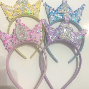 May include: Four colourful sequined crowns with numbers 1, 2, 4, and 6 on them. The crowns are attached to headbands.
