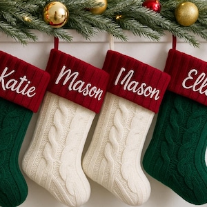 Puede incluir: Cuatro calcetines navideños personalizados cuelgan de una guirnalda. Dos calcetines de punto trenzado verde y dos calcetines de punto trenzado crema están rematados con puños rojos. Cada puño está bordado con un nombre en escritura blanca: Katie, Mason, Mason y Ella.