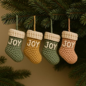 May include: Four miniature crocheted Christmas stockings in shades of green, yellow, and pink, each with a cream-colored cuff and the word "JOY" in white. They are hanging from a green pine branch.