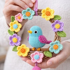 May include: A crocheted wreath featuring a blue bird with pink wings and a yellow beak, surrounded by colorful crocheted flowers. The wreath is adorned with green leaves and a pink bell. The text "CROCHET PATTERN" is at the top.