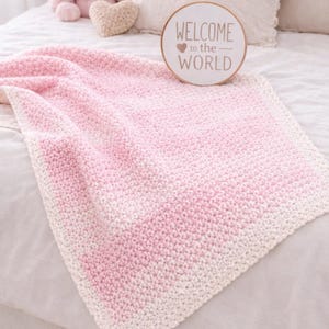 May include: A handmade, pink and white crochet baby blanket. The blanket is folded on a bed with a pink stuffed bunny, a heart-shaped item, and a wooden sign that reads "WELCOME to the WORLD".