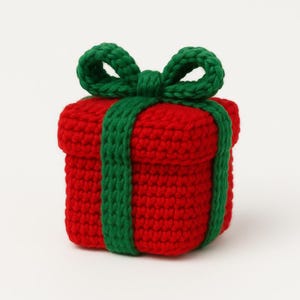 Op de afbeelding: Een handgemaakte, gehaakte geschenkdoos in helder rood, versierd met een groene strik en bijpassend groen lint. De doos is vierkant, met een getextureerd oppervlak gecreëerd door de haaksteken. De strik zit bovenop de doos.