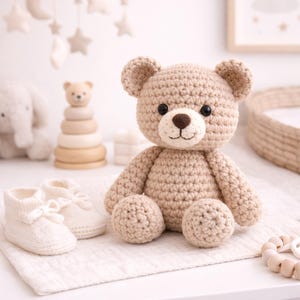 Puede incluir: Un oso de peluche de ganchillo con nariz marrón y ojos negros está sentado sobre una superficie blanca junto a patucos blancos. Un móvil de madera con estrellas y lunas cuelga en el fondo. También se ven otros juguetes para bebés.