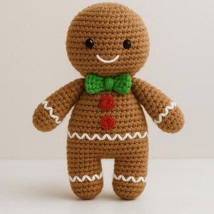 Puede incluir: Muñeco de hombre de jengibre tejido a ganchillo, con cuerpo marrón, ribete en zigzag blanco y pajarita verde. Ojos negros, boca sonriente y dos botones rojos en el pecho. Una decoración navideña festiva.
