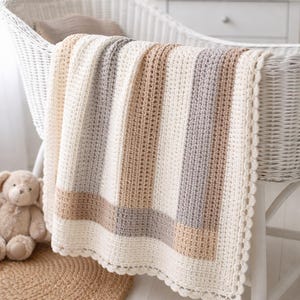 Pode incluir: Um cobertor de bebê listrado em tons neutros de creme, bege e cinza, drapeado sobre um berço de vime branco. O cobertor apresenta uma costura texturizada em forma de waffle e uma borda ondulada. Um ursinho de pelúcia está por perto.