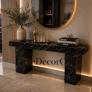 May include: A black marble console table with gold veining featuring a rectangular top and two thick legs. On the table sits a ribbed beige vase with greenery, a gold cocktail shaker, a glass, and a stack of three books labeled Tom Ford, Chanel, and Vogue.
