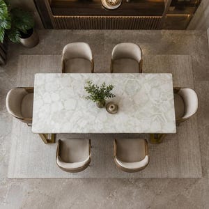 May include: A rectangular dining table with a white stone top featuring a natural veined pattern and gold-toned metal legs, surrounded by six beige upholstered chairs with curved backrests. A small green plant sits in the center of the table.
