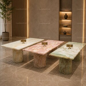 May include: Three rectangular marble tables in cream, pink, and green. Each table has a cylindrical base made of vertical, textured elements. The tables are in a well-lit, modern space.