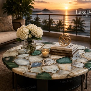 May include: A round coffee table featuring a tabletop composed of polished, multi-colored agate slices in shades of green, white, and grey. The table has a dark base and is topped with a glass vase of white hydrangeas, a candle, and books.