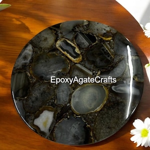 Puede incluir: Una bandeja redonda de ágata negra y blanca con detalles dorados. La bandeja está hecha de resina epoxi y presenta una variedad de rodajas de ágata. La bandeja está sobre una superficie de madera. EpoxyAgateCrafts está impreso en la bandeja.