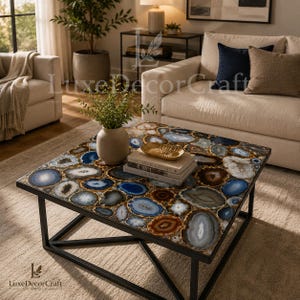 May include: Square coffee table with a unique agate stone top in shades of blue, brown, and white, set in a black metal frame. The table is in a living room setting with a beige sofa and rug. A vase with greenery and books are on the table.