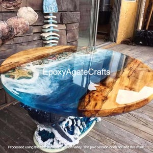 Op de afbeelding: Een ronde houten tafel met een blauw en wit epoxyhars blad dat lijkt op een oceaan golf. De tafelpoot is gebeeldhouwd om te lijken op golven die breken tegen een rotsachtige kust.