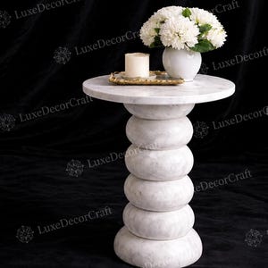 Op de afbeelding: Een bijzettafel van wit marmer met een rond blad en een gestapelde, ronde voet. Op de tafel staat een witte keramische vaas gevuld met witte bloemen, een klein rechthoekig goudkleurig dienblad en een cilindervormige crèmekleurige kaars.