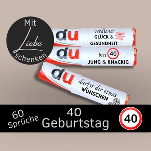 May include: Three red and white tubes with the word "du" printed on them. The tubes have different messages printed on them, including "verdienst GLÜCK & GESUNDHEIT", "bist 40 JUNG & KNACKIG", and "darfst dir etwas WÜNSCHEN". The tubes are arranged in a stack, with the top tube being the most visible. The background is a light brown color. The text "60 Sprüche 40 Geburtstag 40" is printed below the tubes.
