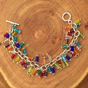 Puede incluir: Un brazalete de cadena de plata con cuentas de vidrio de colores. Las cuentas están dispuestas en forma de cascada y son de varios colores, incluyendo rojo, naranja, amarillo, verde, azul, morado y transparente.