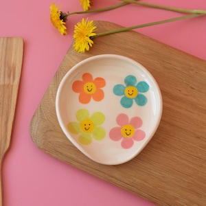 May include: A white ceramic spoon rest with four colorful flower designs, each with a smiling face. The spoon rest sits on a wooden cutting board, with a wooden spatula and yellow flowers in the background. The background is pink.