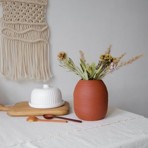 Peut inclure: Un vase en terre cuite de forme ronde et légèrement bombée, rempli de fleurs et d'herbes séchées. Le vase est posé sur une nappe blanche, avec une planche à découper en bois et deux cuillères en bois en arrière-plan.
