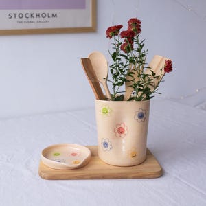 Może przedstawiać: Beżowy ceramiczny pojemnik na przybory kuchenne i podstawka pod łyżkę z kolorowymi uśmiechniętymi wzorami kwiatów. Pojemnik zawiera drewniane łyżki i czerwone kwiaty. Zestaw stoi na drewnianej tacy.