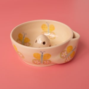 May include: A cream-colored ceramic bowl featuring a repeating pattern of yellow and pink butterflies on the exterior and interior. The bowl includes a central, raised, dome-shaped element with small circular perforations and a side spout.